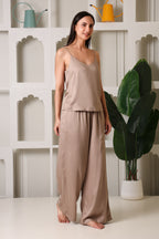 A woman modeling a two-piece lounge set in a warm taupe-beige color, consisting of a thin-strap camisole top and matching wide-leg pants, standing in front of a white decorative wall with shelves and a potted plant.